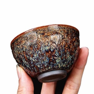 Puede incluir: Un pequeño cuenco de cerámica con una superficie texturizada y un esmalte moteado en tonos marrones, azules y dorados. El cuenco tiene forma redondeada, un borde acampanado y una base gris oscuro. El cuenco se sostiene en una mano.