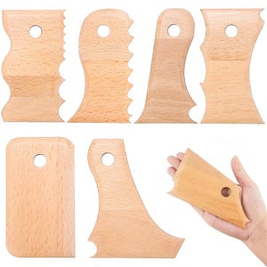 Könnte beinhalten: Sieben Holz-Töpferwerkzeuge, darunter verschiedene geformte Rippen und ein rechteckiges Glättwerkzeug. Jedes Werkzeug ist hellbraun mit glatter Oberfläche und einem Loch zum Aufhängen. Ein Werkzeug wird in der Hand gehalten, um Größe und Form zu zeigen.