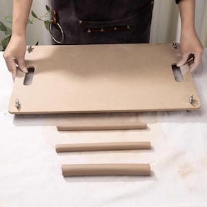 Könnte beinhalten: Eine hellbraune Holz-Tontafel mit zwei Griffaussparungen und Metallbefestigungen. Drei gerollte Tonstücke befinden sich unter der Platte. Die Gegenstände befinden sich auf einer weißen Oberfläche. Die Person trägt eine braune Schürze.