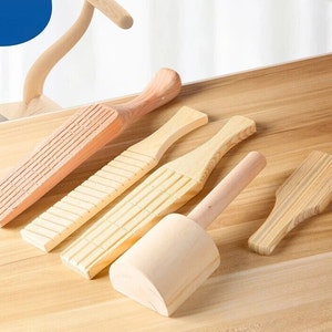 May include: Five wooden kitchen tools, including a rolling pin, a dough scraper, and three different types of pasta cutters. The tools are made of light brown wood and have a natural finish.