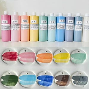 May include: A collection of colorful art supplies, including bottles of liquid paint in various shades like pink, yellow, and blue, and ceramic color swatches. The swatches display painted strokes of the corresponding colors, offering a visual guide.
