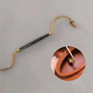May include: A brass-colored, curved tool with a black handle and pointed tip. The tool has a hook shape on one end and a conical tip on the other. A close-up shows the tool's use.