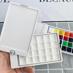 以下が含まれることがあります： 白いプラスチック製の水彩絵の具パレット、ヒンジ付きの蓋付き。 パレットは開いており、空のウェルとさまざまな色の絵の具で満たされた別のパレットが表示されます。 パレットは人の手で持たれています。