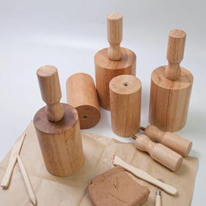 May include: A selection of wooden pottery tools, including cylindrical molds with handles, small rolling pins, and clay sculpting tools. The molds are labeled with measurements in inches. The tools are arranged on a sheet of brown paper.