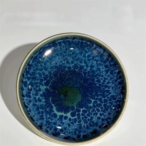 May include: A round ceramic plate with a deep blue and green speckled glaze. The plate has a light beige rim and a glossy finish. The glaze pattern resembles a close-up view of the ocean. This decorative plate is suitable for serving food or as a decorative item.