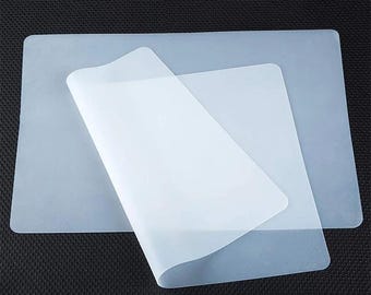 Può includere: Una pila di tappetini in silicone traslucidi e rettangolari. Un tappetino è parzialmente staccato, rivelando un secondo tappetino sottostante. I tappetini sono di un colore pallido e semitrasparente e i bordi sono arrotondati. Lo sfondo è una superficie scura e strutturata.