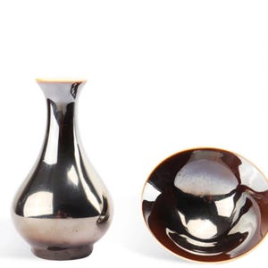 Könnte beinhalten: Zwei Keramikvasen mit glänzendem, metallischem schwarzen Überzug. Die Vase links ist eine traditionelle, schmalhalsige, birnenförmige Vase. Die Vase rechts ist eine flache, schalenförmige Vase.