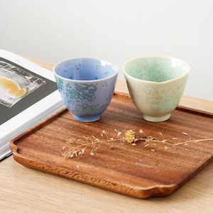 Puede incluir: Dos tazas de té de cerámica, una azul y otra verde, con un patrón moteado, están sobre una bandeja de madera. La taza azul tiene una base azul claro y la taza verde tiene una base color crema. Un libro está abierto en el fondo.