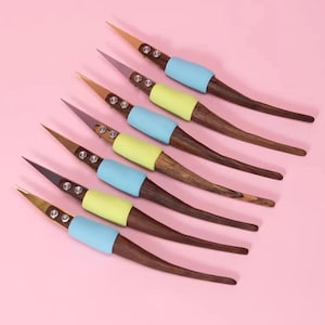 May include: Seven wooden hair sticks with a pointed end and a curved handle are arranged diagonally on a pink background. Each stick has a different colored tip, including gold, brown, and gray. The sticks have a light blue and yellow band.