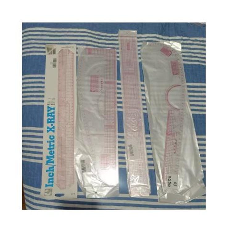 May include: A collection of pink and clear plastic rulers and templates, including a straight ruler, a curved ruler, and a French curve, on a blue and white striped surface. One ruler is labeled "Inch/Metric X-Ray".