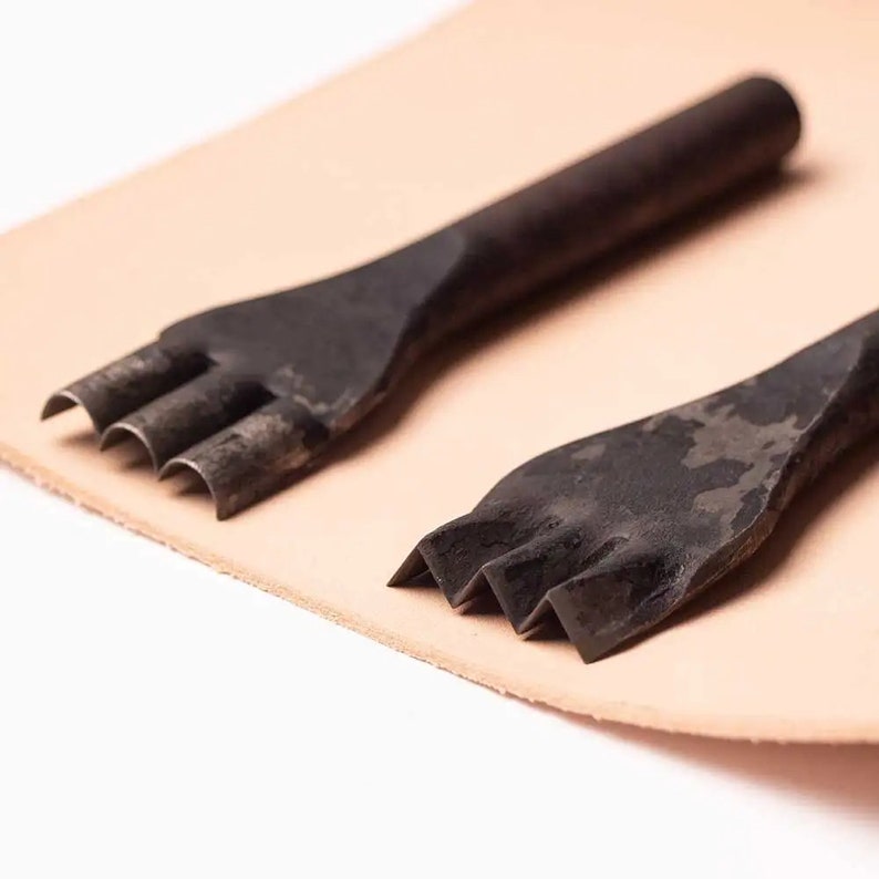 May include: Two black metal leather punches with three prongs each, lying on a piece of tan leather.