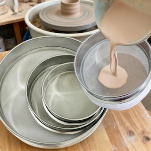 May include: A set of three circular stainless steel sieves with fine mesh screens nested together. A light peach-coloured liquid is being poured from a container through the mesh of the smallest sieve, creating a smooth stream into the centre.