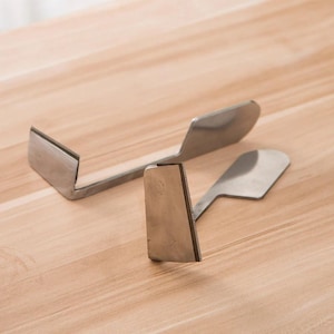 May include: Two stainless steel tools, likely for woodworking or crafting, are displayed on a light wood surface. One tool is L-shaped with a flat blade, and the other has a curved handle and a rectangular blade with a serrated edge. The tools are angled towards the viewer.