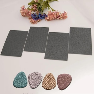 May include: Four rectangular dark gray silicone texture mats featuring intricate patterns of shells, geometric shapes, mushrooms, and leaves, accompanied by four teardrop-shaped clay pieces displaying matching embossed designs.