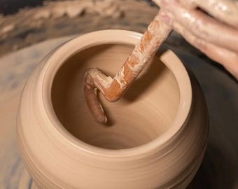 May include: A potter's hands shaping a light beige clay pot on a spinning wheel. The tool is used to refine the inside of the vessel, creating a smooth, rounded form. The pottery process is captured in a close-up shot.