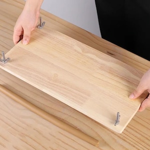 Peut inclure: Une étagère en bois de couleur claire avec des fixations métalliques est tenue par des mains. Une cheville en bois cylindrique est posée à proximité. L'étagère est rectangulaire et semble inachevée.