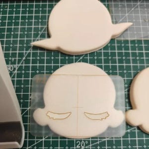 Puede incluir: Primer plano de varias galletas pálidas con forma de fantasma sobre una esterilla de corte verde. Una galleta tiene detalles dorados en forma de ojos y está parcialmente cubierta por una plantilla transparente. Las galletas están dispuestas cerca de un objeto blanco y un transportador.