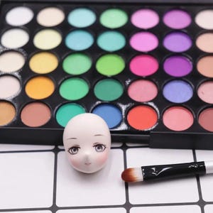 May include: A black makeup palette filled with a spectrum of eyeshadow colours, from pastels to vibrant hues. A doll head with painted facial features and a makeup brush are also visible, indicating a beauty or craft focus.