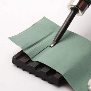 May include: A close-up of a leather working tool being sharpened on a piece of sandpaper. The tool has a small, pointed blade and is held in a metal holder. The sandpaper is placed on a black, textured surface.
