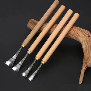 May include: Four wood carving tools with long, cylindrical light-colored wooden handles and brass-toned metal ferrules. Each tool features a distinctively shaped, polished metal blade with a flared, flat edge, arranged in a descending size order.