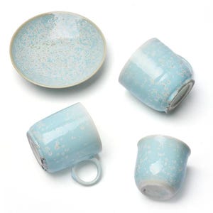May include: A ceramic tea set consisting of one saucer and three mugs, all featuring a light blue glaze with a white speckled pattern. The mugs have rounded bodies and small handles, while the saucer has a wide, shallow, circular shape.
