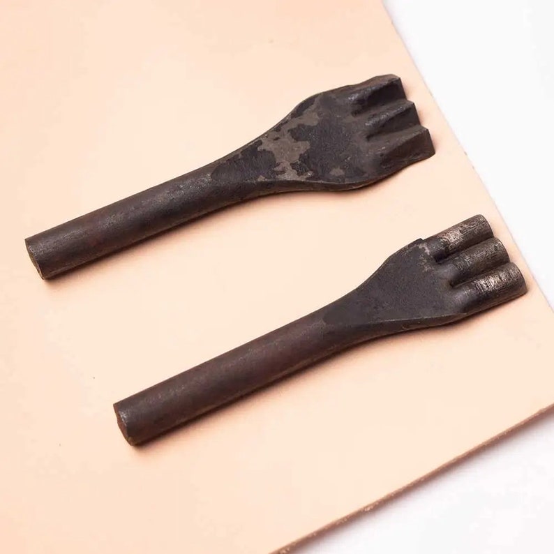 May include: Two metal leather punches with three prongs each, lying on a light brown leather surface.