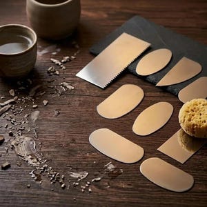 May include: A collection of metal pottery ribs and scrapers in various shapes, including rectangular, oval, and serrated edges, alongside a small natural yellow sponge. These tools are arranged on a dark slate board and a wooden surface with ceramic cups.