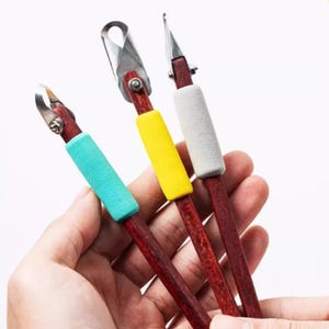 Pode incluir: Três ferramentas para esculpir argila com cabos de madeira castanho-avermelhada escura e pontas de metal. Cada ferramenta possui um punho de espuma macia de cor diferente: turquesa, amarelo e cinza claro. As pontas de metal variam em formato.