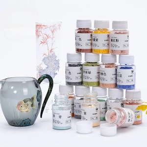 Puede incluir: Una colección de polvos pigmentados de colores en pequeños frascos transparentes con tapas blancas. Los frascos están apilados cerca de una jarra de vidrio con un diseño de pez dorado y un vaso alto con un patrón floral. Los polvos están etiquetados con caracteres chinos.