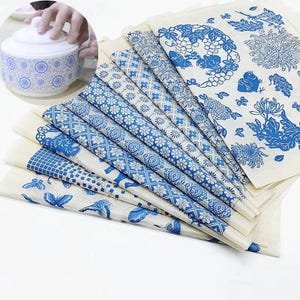 Könnte beinhalten: Ein Stapel rechteckiger Stoffbögen mit verschiedenen blau-weißen Mustern, darunter Blumen-, Schmetterlings- und geometrische Designs. Ein kreisförmiger Einsatz zeigt eine weiße Keramikschale mit blauem Blumenmuster, die von einer Hand gehalten wird.