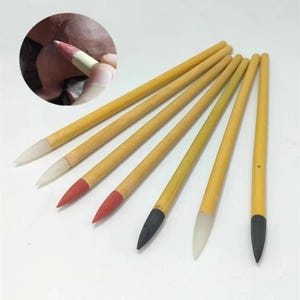 May include: A collection of calligraphy brushes with light brown wooden handles and various tip colors, including red, black, and white. The tips appear to be made of a soft material. A close-up shows a brush in use.