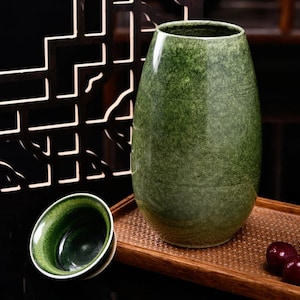 May include: A green ceramic vase with a textured surface and a wide opening. A small, matching green ceramic cup is next to the vase. The objects are on a wooden tray, with a dark background.