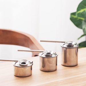 Op de afbeelding: Drie zilveren metalen theepotten met tuiten en deksels. De theepotten zijn gerangschikt in een rij van de grootste tot de kleinste op een houten oppervlak.