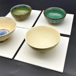 Peut inclure: Quatre petits bols en céramique disposés sur des carreaux carrés blancs individuels. La collection comprend un bol vert avec une base texturée, un bol turquoise vif, un bol crème avec un centre bleu et un bol crème uni.