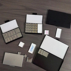 Puede incluir: Un surtido de pequeñas cajas rectangulares de metal y negras, algunas abiertas para revelar cuadrados plateados y papel blanco. Varios pequeños trozos de metal cuadrados y piezas blancas rectangulares están esparcidos alrededor de las cajas. Algunas cajas están abiertas.