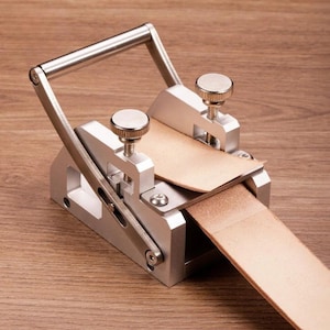 May include: A silver-coloured metal leather skiving tool featuring a sturdy base, an adjustable handle, and two knurled tension knobs. A strip of light tan leather is fed through the mechanism, which is designed to thin the material for crafting.