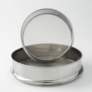 Może przedstawiać: Dwa sita ze stali nierdzewnej, jedno na drugim. Mają drobną siatkę i błyszczące, metaliczne wykończenie. Okrągła konstrukcja nadaje się do przesiewania suchych składników lub przecedzania płynów w kuchni.