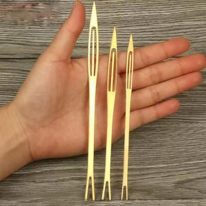 May include: Three gold-colored metal netting needles of varying lengths, each featuring a pointed tip, a long central slot for thread, and a split-end base. These slender, flat tools are designed for weaving or repairing mesh and netting materials.
