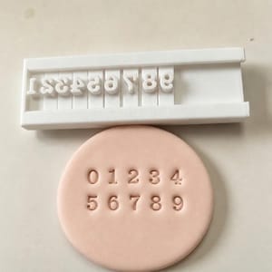 May include: A white plastic number stamp set featuring individual characters from zero to nine arranged in a rectangular holder, positioned above a circular light peach-colored clay disc embossed with the numbers zero through nine.