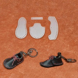 Pode incluir: Um conjunto de chaveiros de sapatos de bebê de couro preto com costuras e cadarços rosa. Também são apresentados moldes de sapatos de couro branco com furos perfurados.