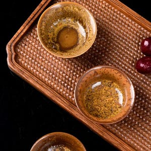 Peut inclure: Trois tasses à thé en céramique ronde avec un vernis moucheté brun et doré, disposées sur un plateau en bois rectangulaire au motif tressé. Deux fruits ronds rouge foncé sont placés sur le côté droit de la surface du plateau.