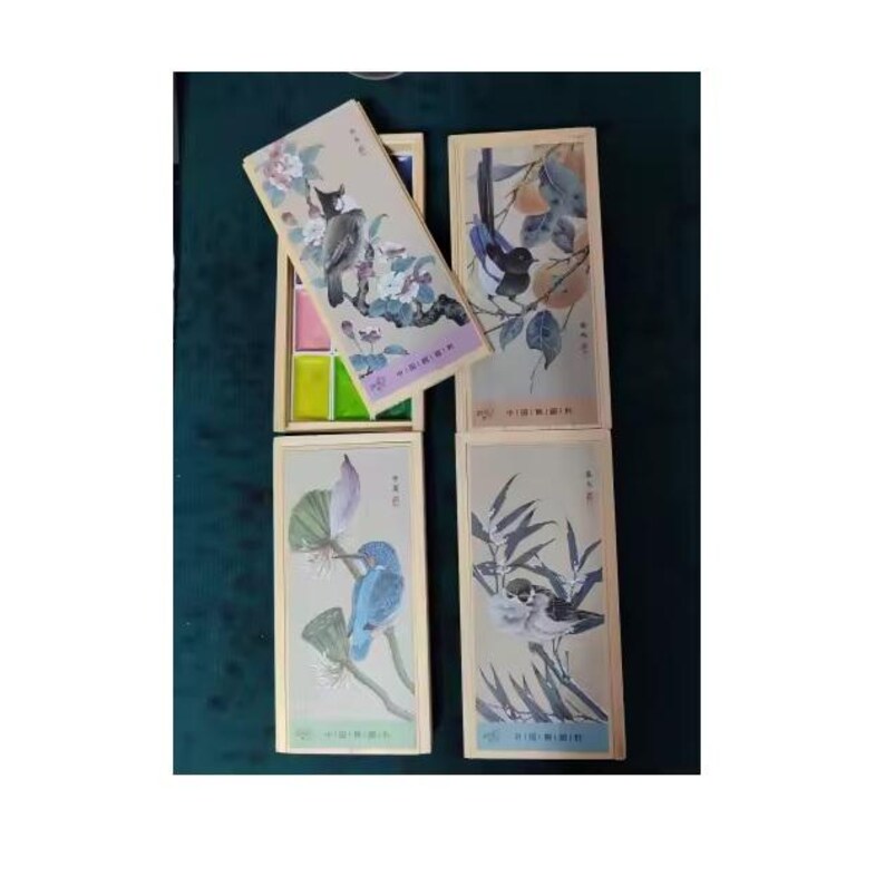 May include: Watercolor paint set with bird illustrations. The set includes a wooden box with watercolor pans and several rectangular boxes with bird and floral designs. The bird illustrations are in shades of blue, green, and gray.