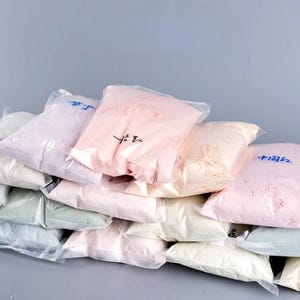 May include: A collection of clear plastic bags filled with fine, powdery substances in various pastel shades including light pink, pale green, and off-white. Some bags feature handwritten Chinese characters in blue or black ink on the exterior surface.