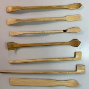 May include: A collection of seven light brown bamboo tools arranged horizontally. The set includes various shapes such as flat spatulas, pointed ends, and L-shaped implements, all featuring a natural wood grain texture and smooth, finished surfaces.