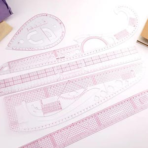 May include: A collection of clear, pink-tinted plastic sewing rulers and templates. Various shapes include straight rulers, curved rulers, and a teardrop-shaped template. The rulers have measurement markings in inches and centimeters.