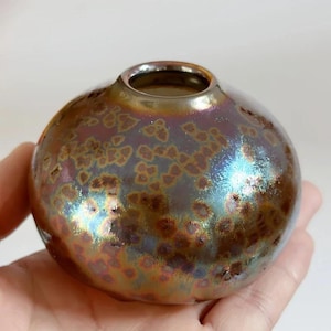 Puede incluir: Un pequeño jarrón de cerámica redondo con un esmalte metálico iridiscente. El jarrón tiene una abertura ancha y un cuerpo bulboso. El esmalte muestra tonos de oro, bronce y azul. El jarrón se sostiene en una mano.