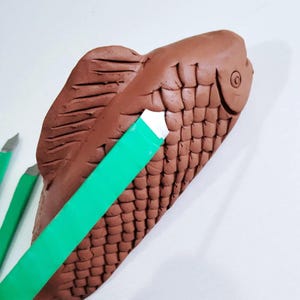 Könnte beinhalten: Eine braune Fischskulptur aus Ton mit detaillierten Schuppen und Flossen. Ein grünes Bildhauwerkzeug liegt über dem Fischkörper, mit zwei weiteren Werkzeugen im Hintergrund. Das Kunstwerk ist vor einem weißen Hintergrund.
