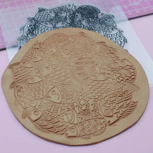 May include: A brown rubber stamp with a design of multiple fish with intricate patterns. The stamp is circular and has a raised design.