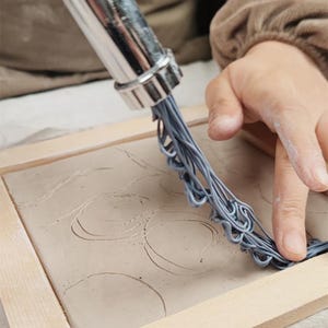 Puede incluir: Primer plano de un proyecto de arte en arcilla en progreso. Una herramienta plateada extruye hebras de arcilla gris sobre una base rectangular de arcilla dentro de un marco de madera. Se ve la mano del artista, guiando la herramienta para crear un diseño texturizado.