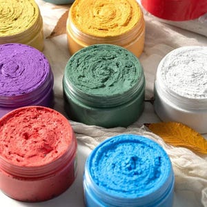May include: A collection of round plastic jars filled with thick, textured cosmetic paste in various colors including yellow, green, purple, red, blue, and white. Each jar features a wide opening revealing the swirled, dense consistency of the product.